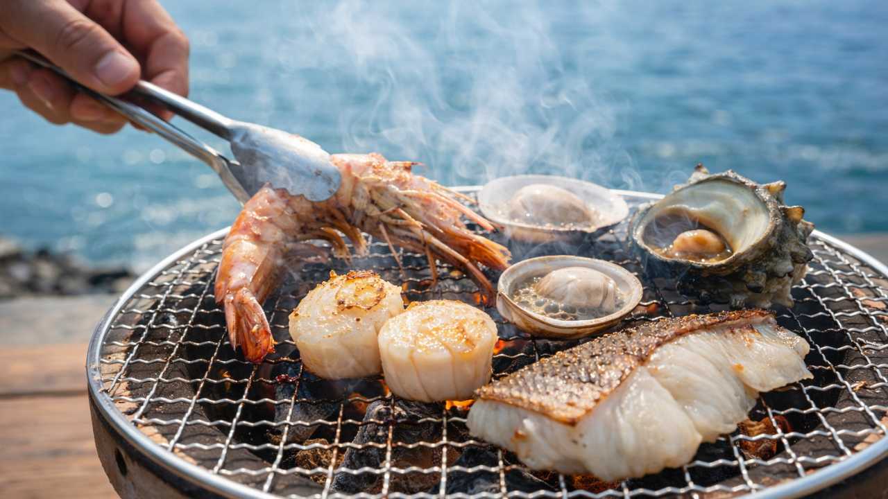 七輪の上で魚介（貝・エビ・白身魚）を焼いている手元のクローズアップ