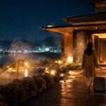 広島の夜景を背景に、温かい湯けむりが立ちのぼるモダンな温浴施設の外観、旅人がスーツケースを持って入口へ向かう、清潔感のある実写風