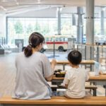 博物館の1階休憩スペースをイメージした明るいベンチと親子のお弁当ランチ、清潔で落ち着いた写真風